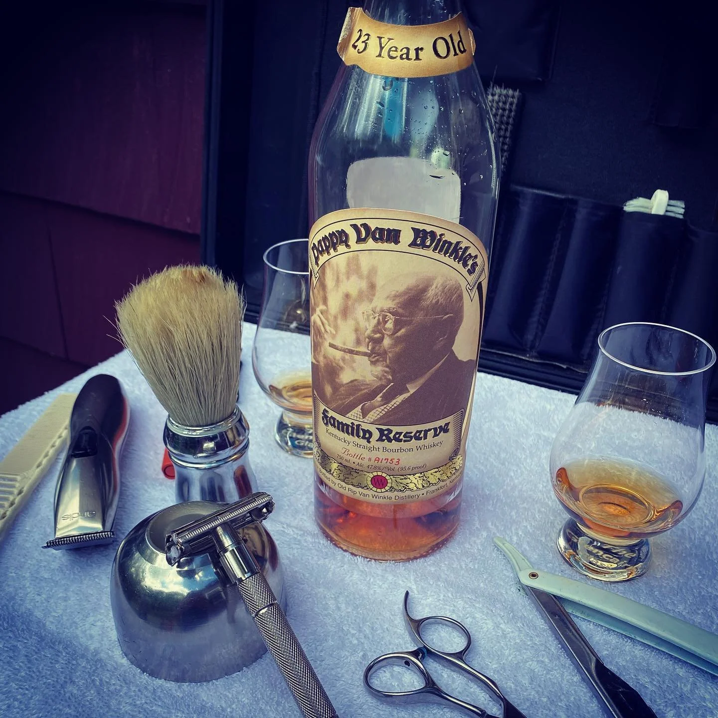 Pappy Van Winkle 23 Year Old bourbon bottle with barbershop tools and whiskey glasses on a towel.