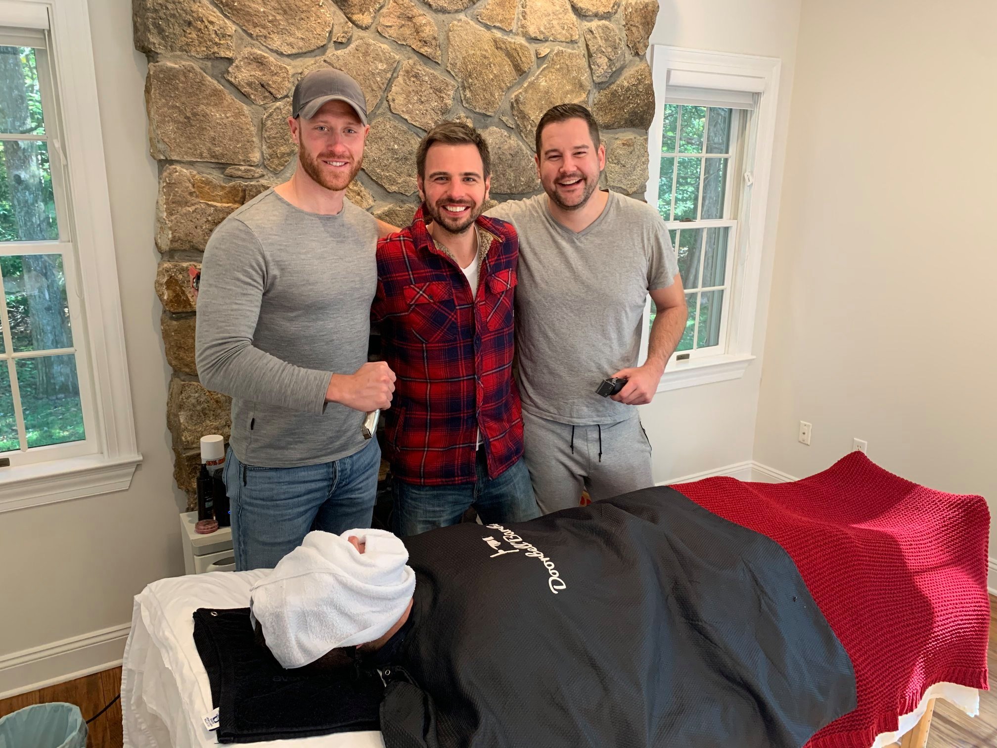 Three men smiling and standing indoors next to a massage table with a person lying down under a towel, with a stone wall and windows in the background.