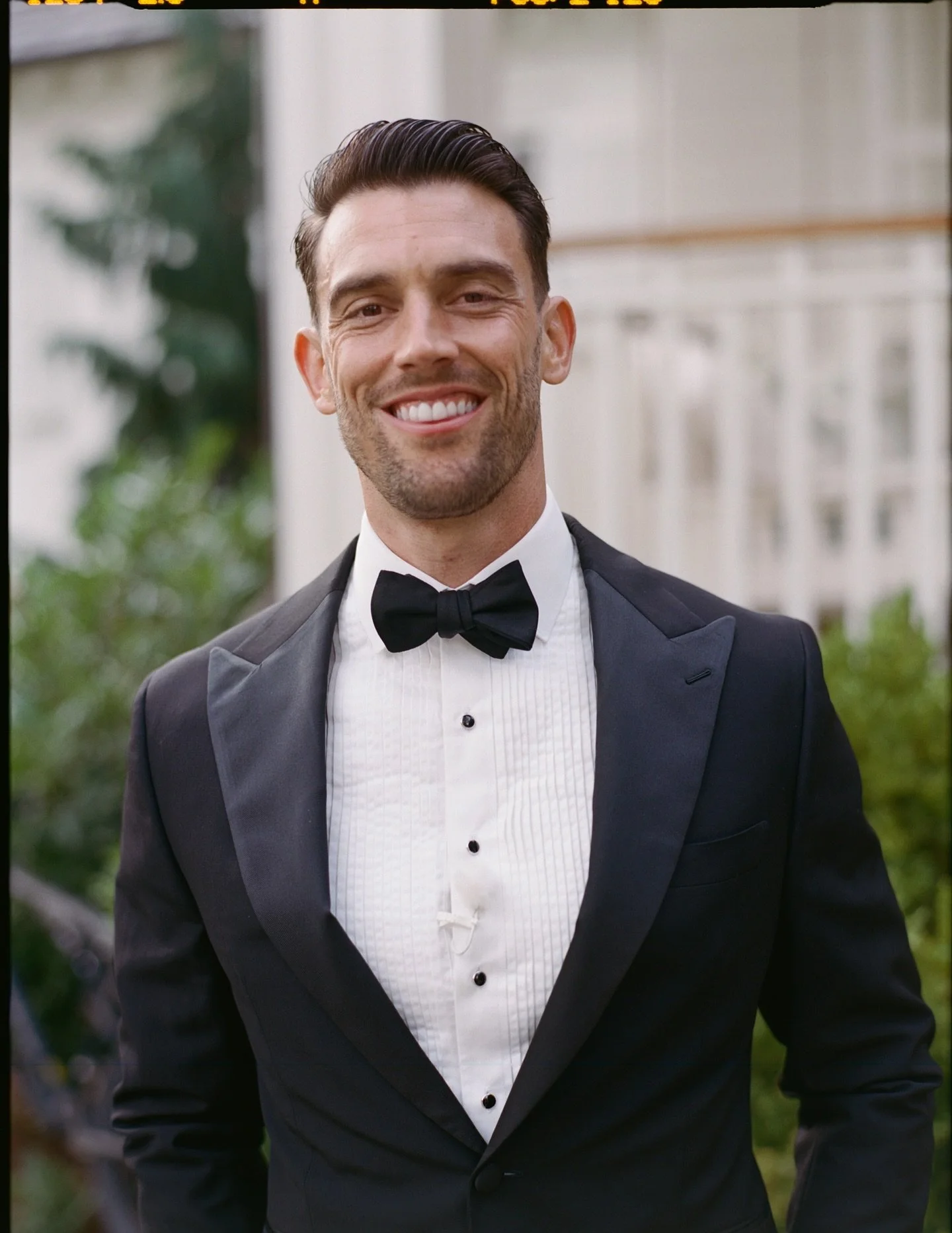 A man in a black tuxedo with a bow tie, smiling outdoors with a blurred architectural and greenery background.