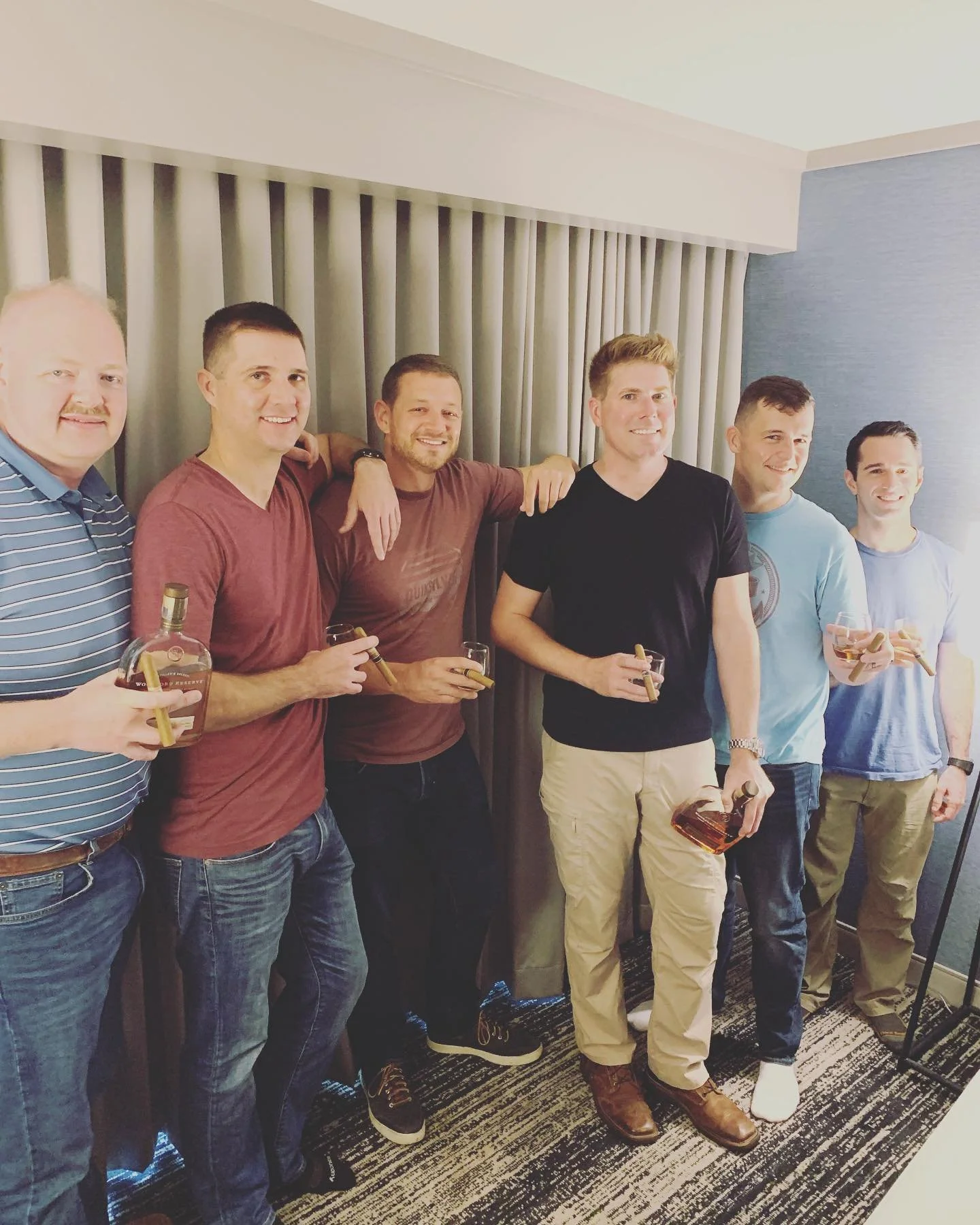 Group of six men holding cigars and drinks, standing together in a room with a curtain backdrop.