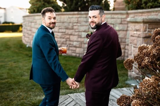 Two men in formal suits holding hands and smiling outdoors, near a stone wall and dry plants.