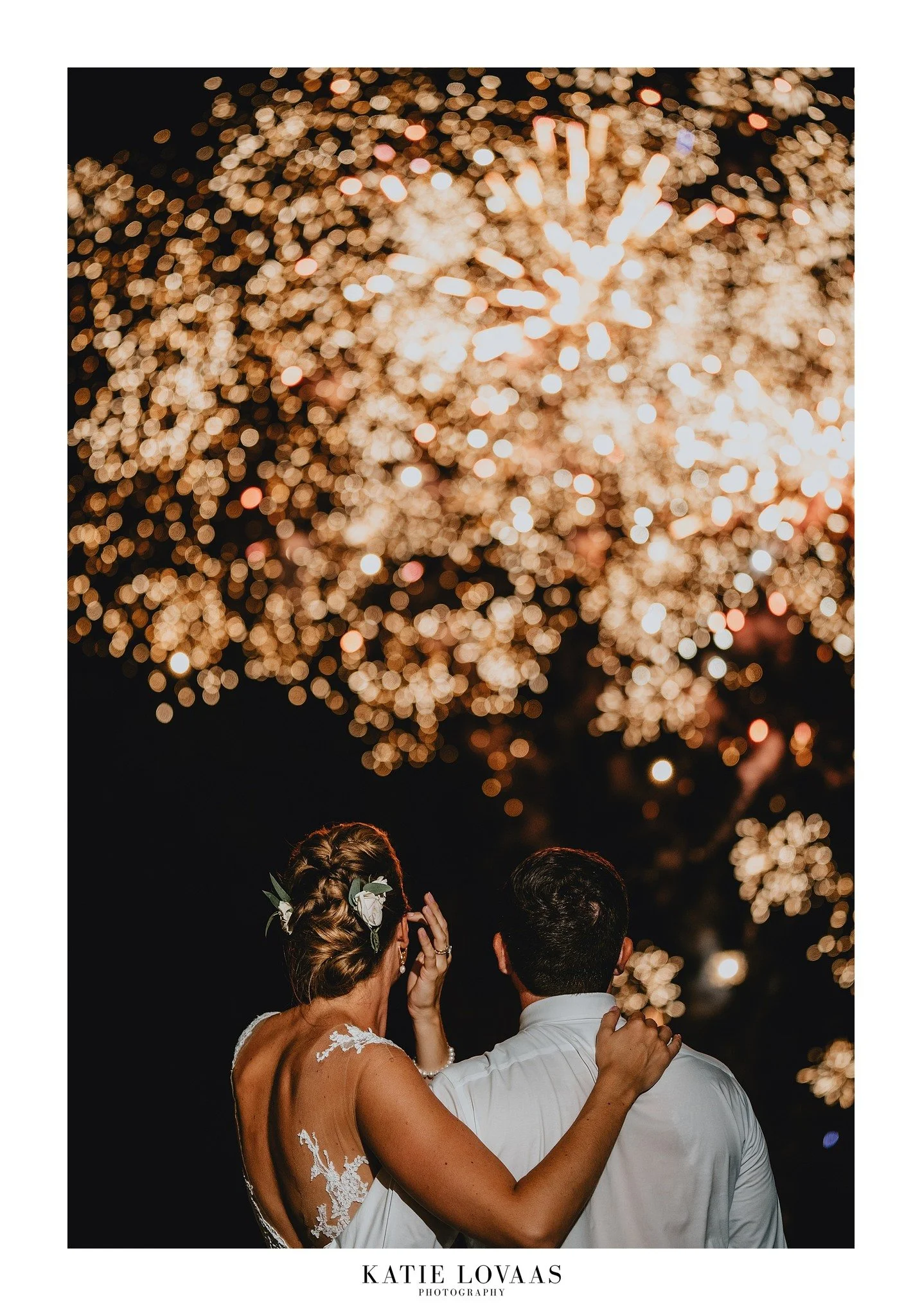 Couple watching fireworks display at night, with bride in lace dress embracing her partner.