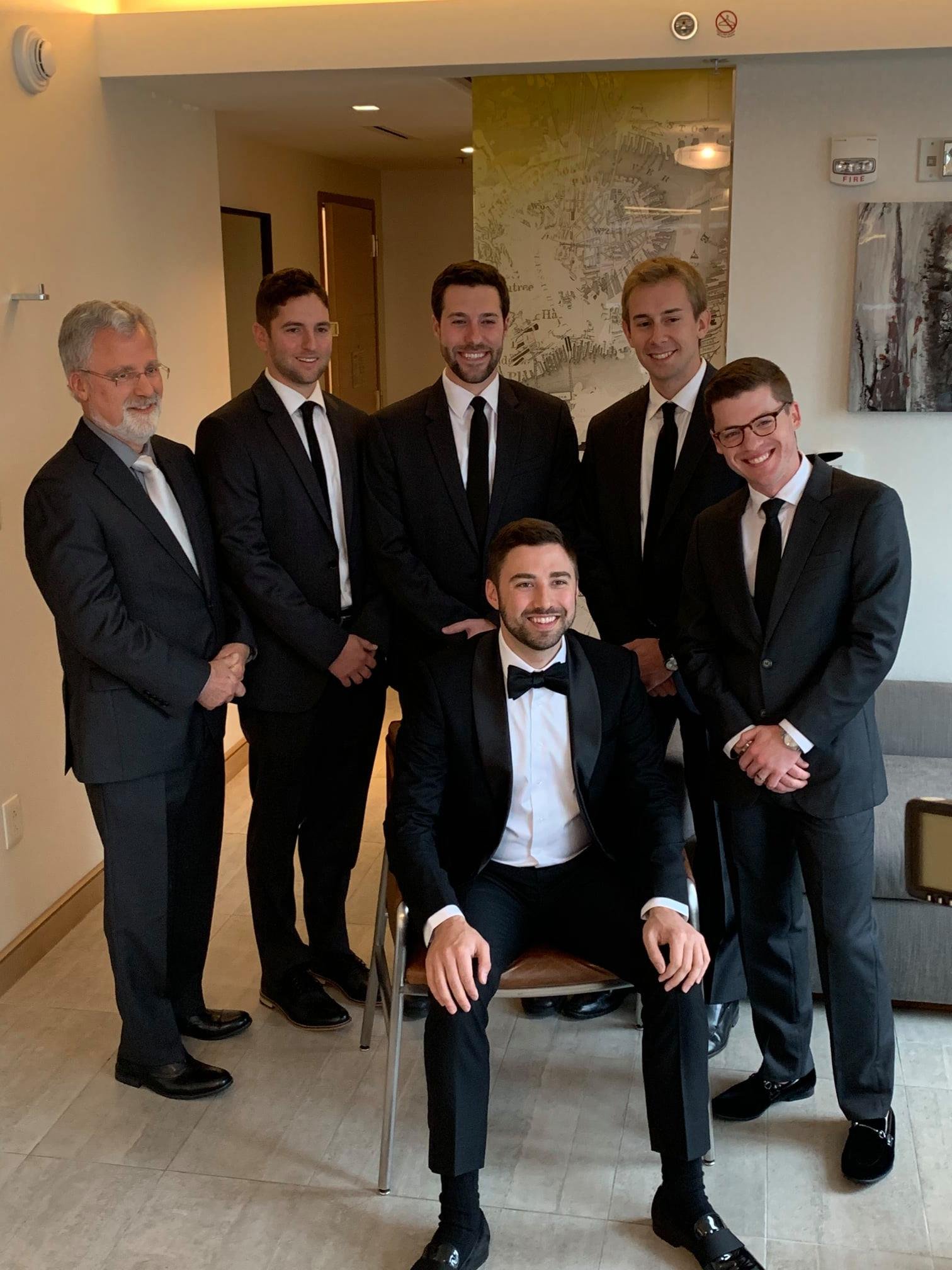 Group of six men in suits posing indoors, one seated and smiling, in a modern room.