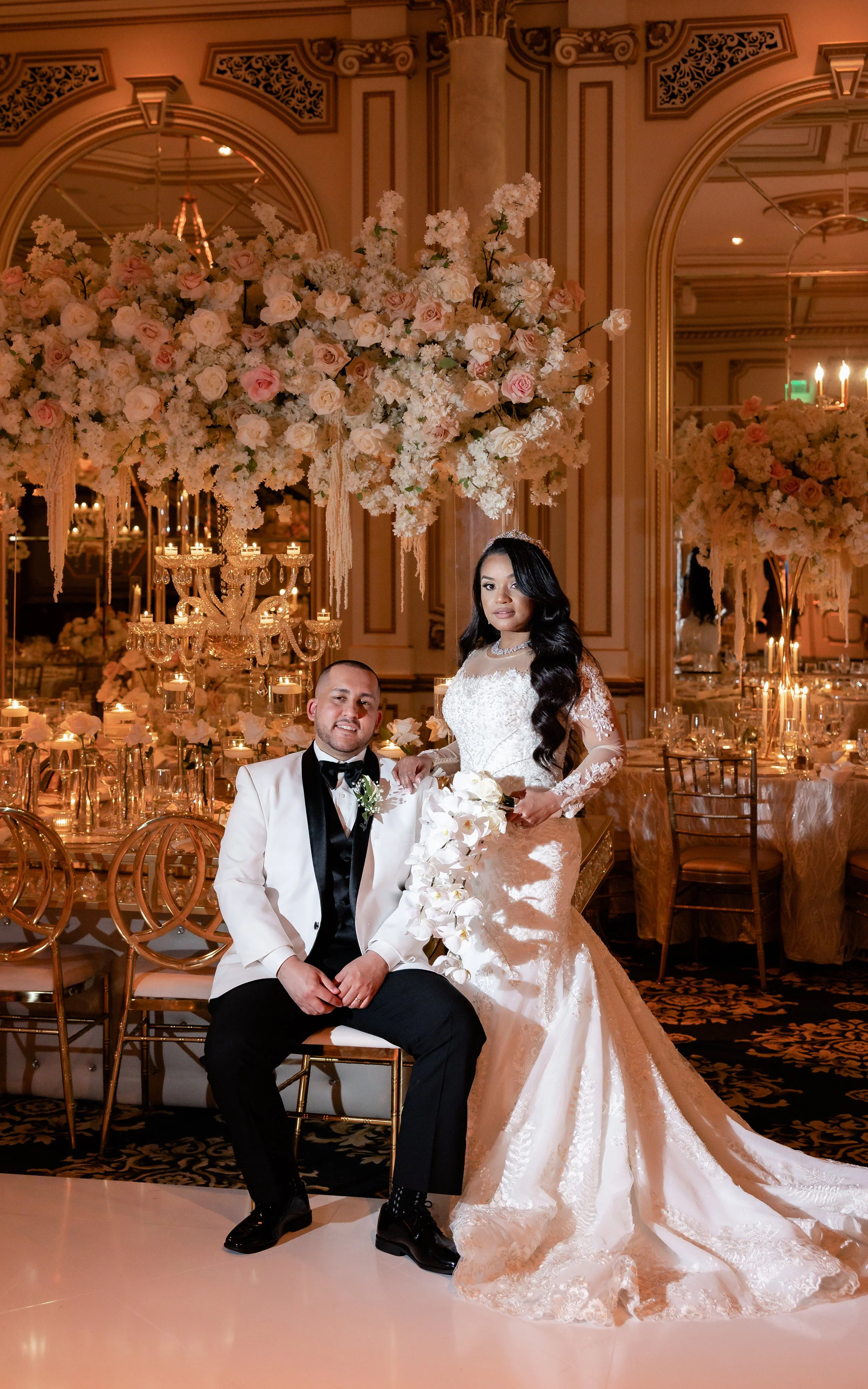 Bride and groom in formal attire at a lavish wedding venue with floral arrangements and chandeliers.