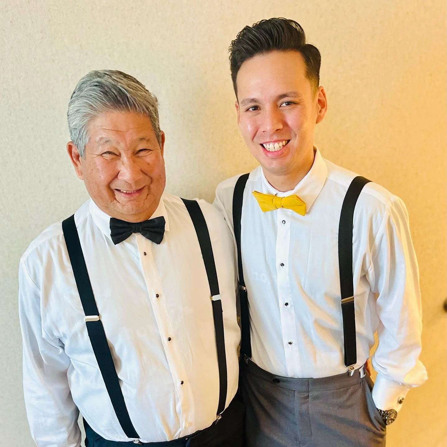 Two men wearing white dress shirts and suspenders, one with a black bow tie and the other with a yellow bow tie, standing side by side and smiling.