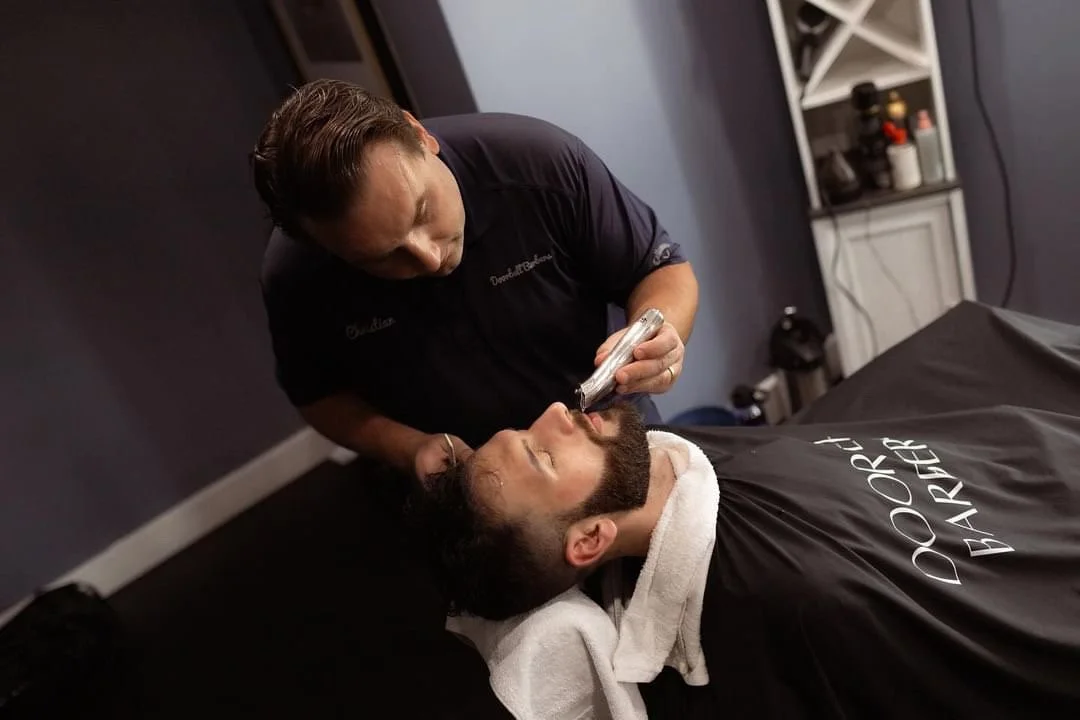 Barber trimming a man's beard with clippers in a barbershop.