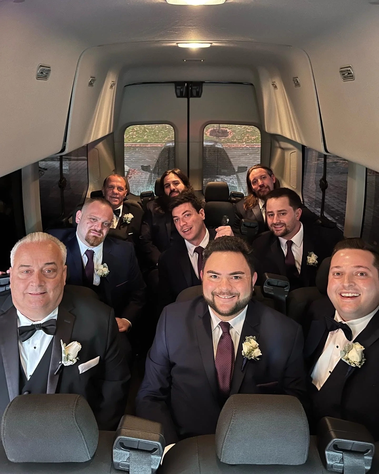 A group of men dressed in formal suits with boutonnieres, seated inside a van, smiling for the photo. They appear to be part of a wedding party, possibly groomsmen.