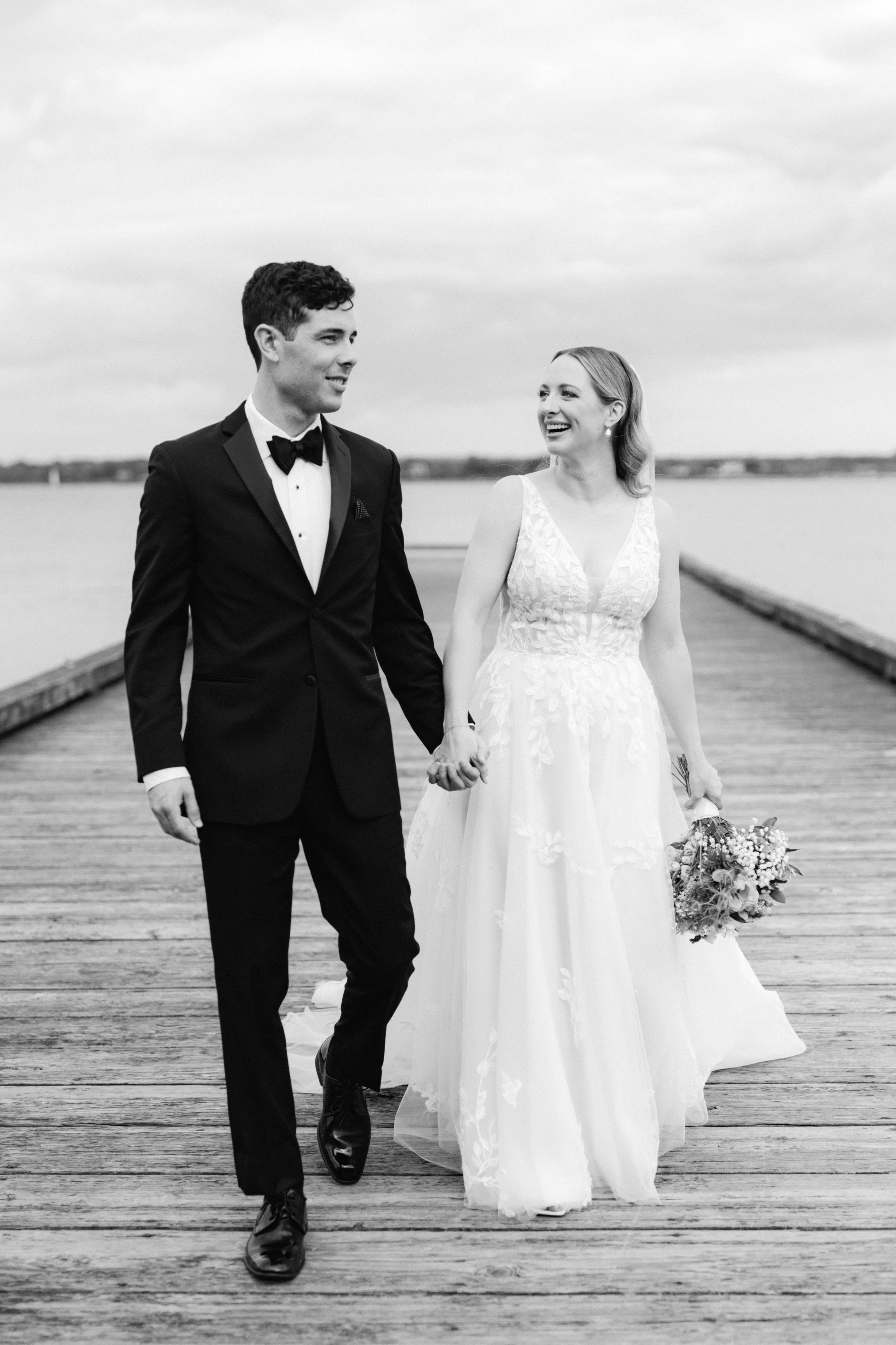Bride and groom walking on a wooden dock, holding hands, with water in the background.