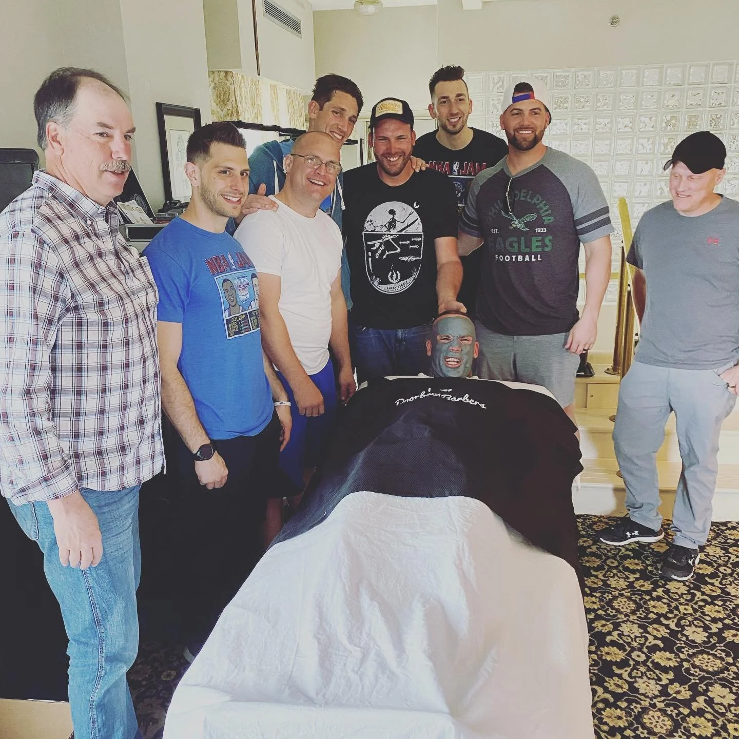 Group of men standing around a person lying on a table with a face mask, smiling indoors.