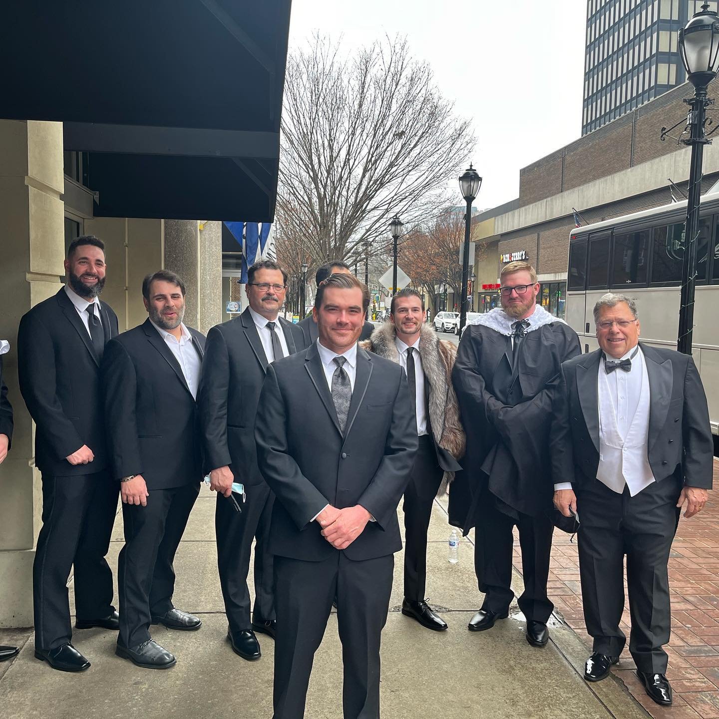 Group of men in suits on a city sidewalk, some wearing overcoats and one with a cape, near a bus.