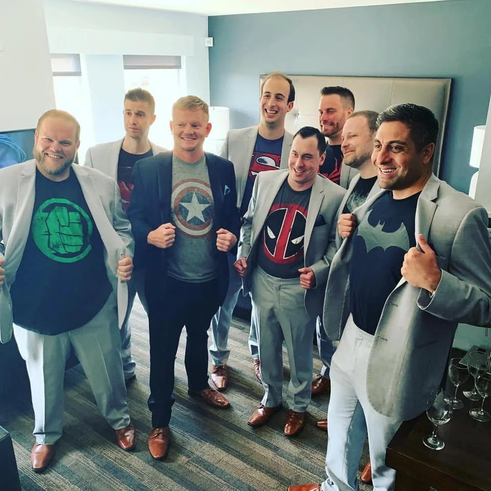 Group of men in suits wearing superhero t-shirts, smiling and posing indoors.
