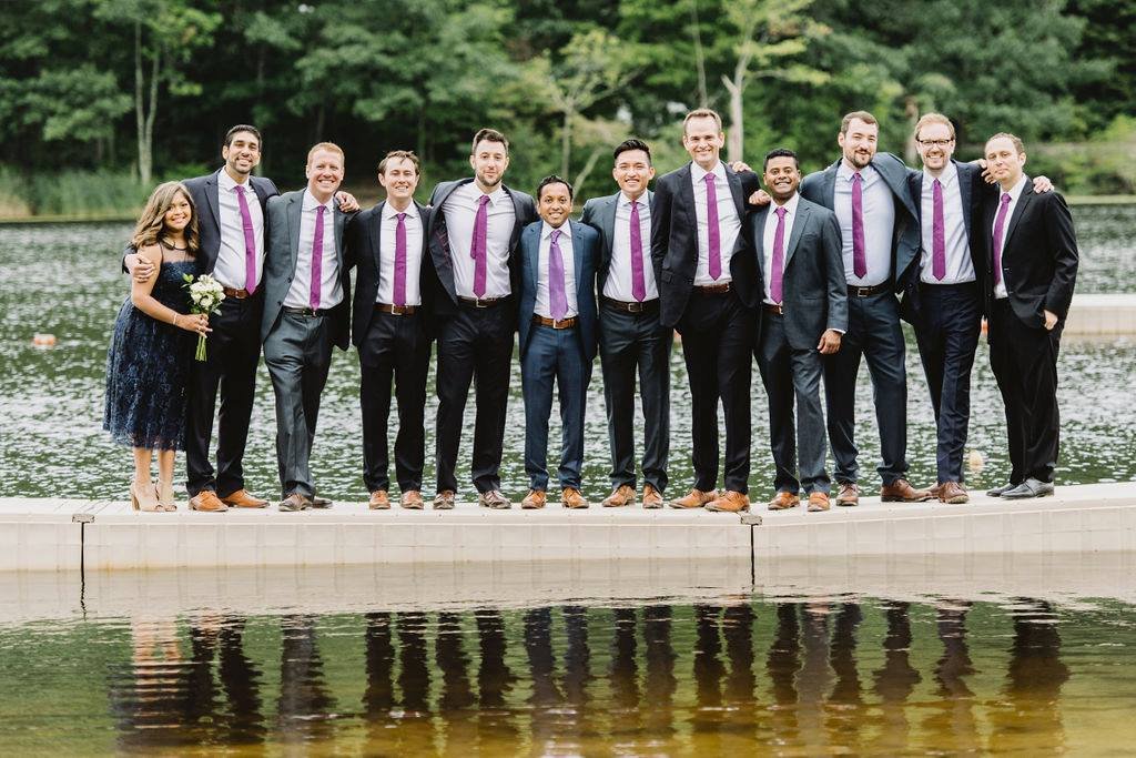 A group of men in suits with matching purple ties, standing next to a woman in a blue dress holding flowers by a lakeside.