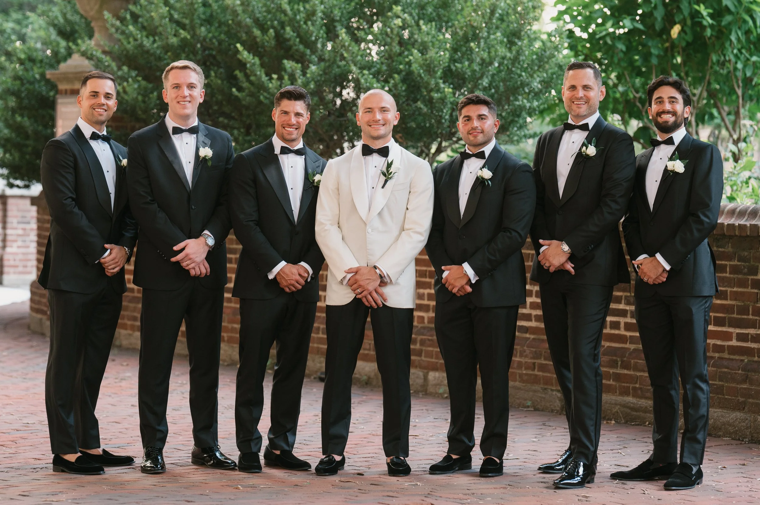 A group of seven men dressed in tuxedos, standing outdoors on a brick path with greenery and trees in the background. The man in the middle is wearing a white tuxedo jacket, while the others are in black tuxedos. All are smiling and have boutonnieres