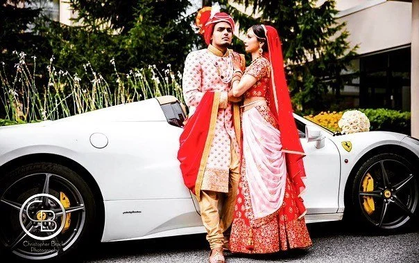 A couple in traditional Indian wedding attire stands beside a white luxury car, outdoors, with greenery in the background.
