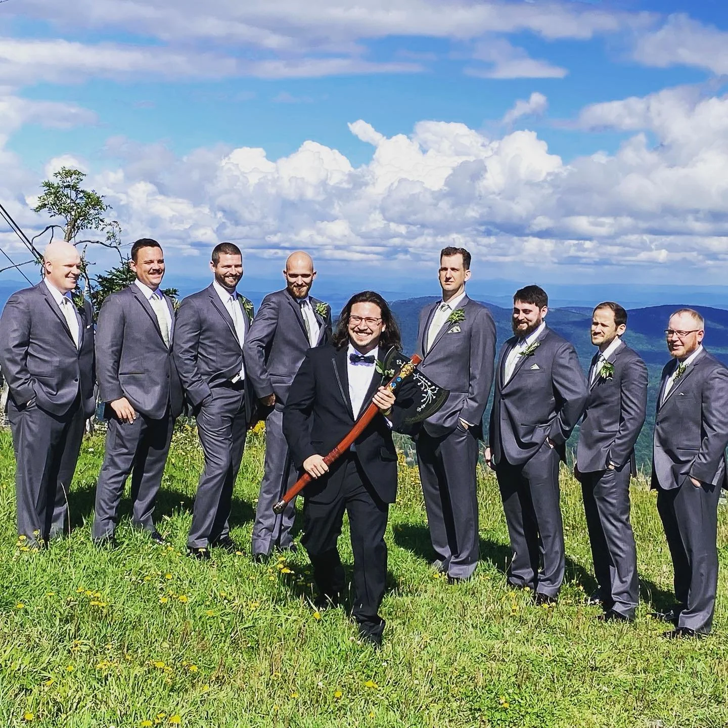 Nine men in suits standing on a grassy hillside with a blue sky and clouds, one holding an ornate axe.
