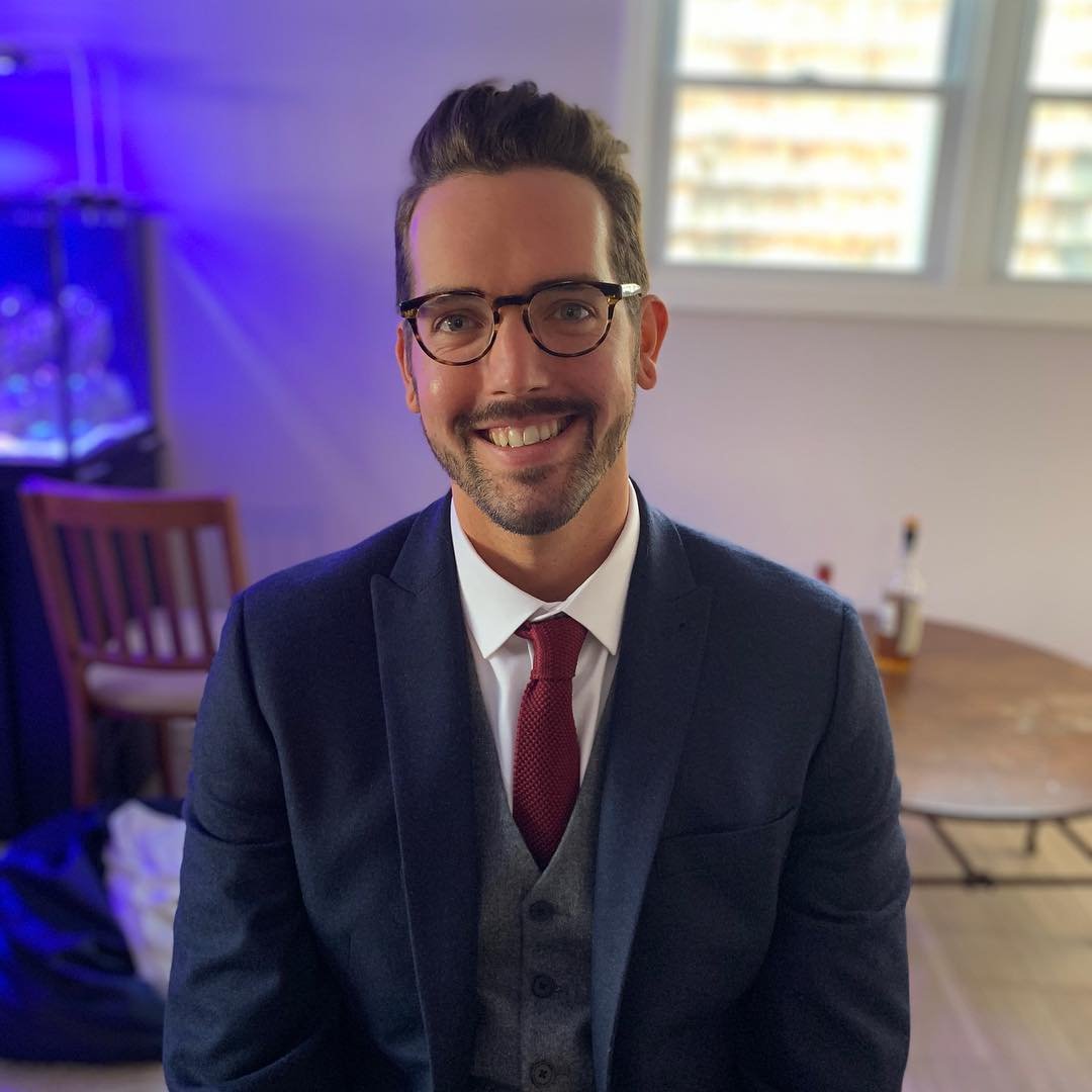 Smiling man wearing glasses and a suit with a red tie, sitting indoors with a blurred background including a chair and bottles on a table.