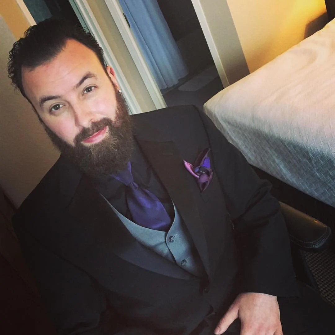 Man in formal attire with a beard, wearing a dark suit, purple tie and pocket square, sitting indoors.