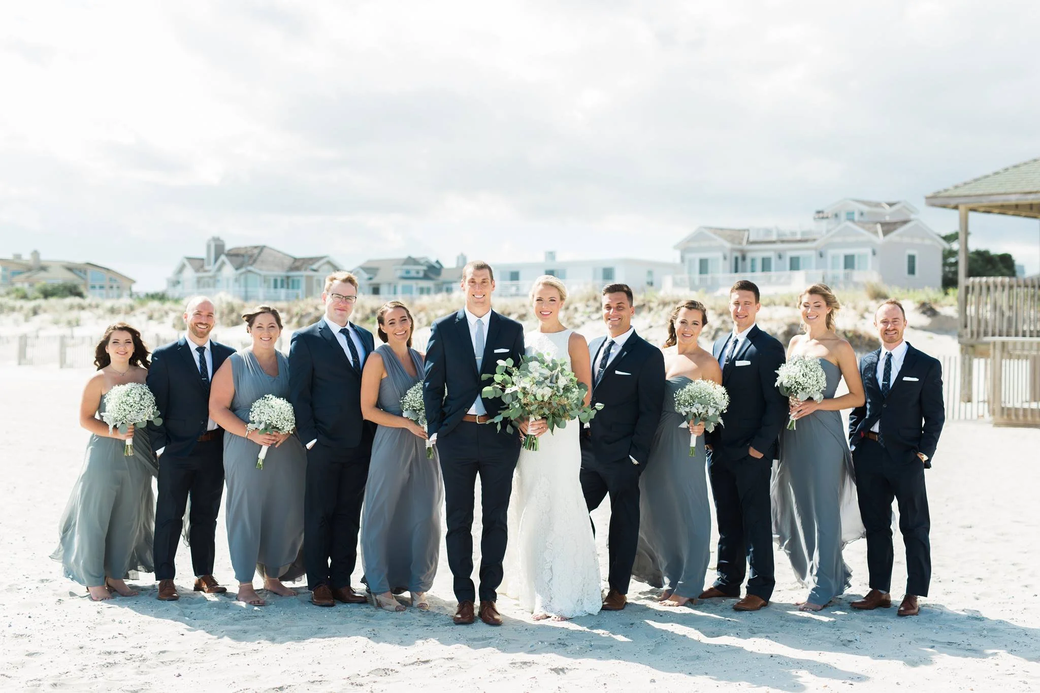 A wedding party posing on a beach, with the bride and groom in the center surrounded by bridesmaids in gray dresses and groomsmen in dark suits. The group is smiling, holding flowers, with beach houses in the background.