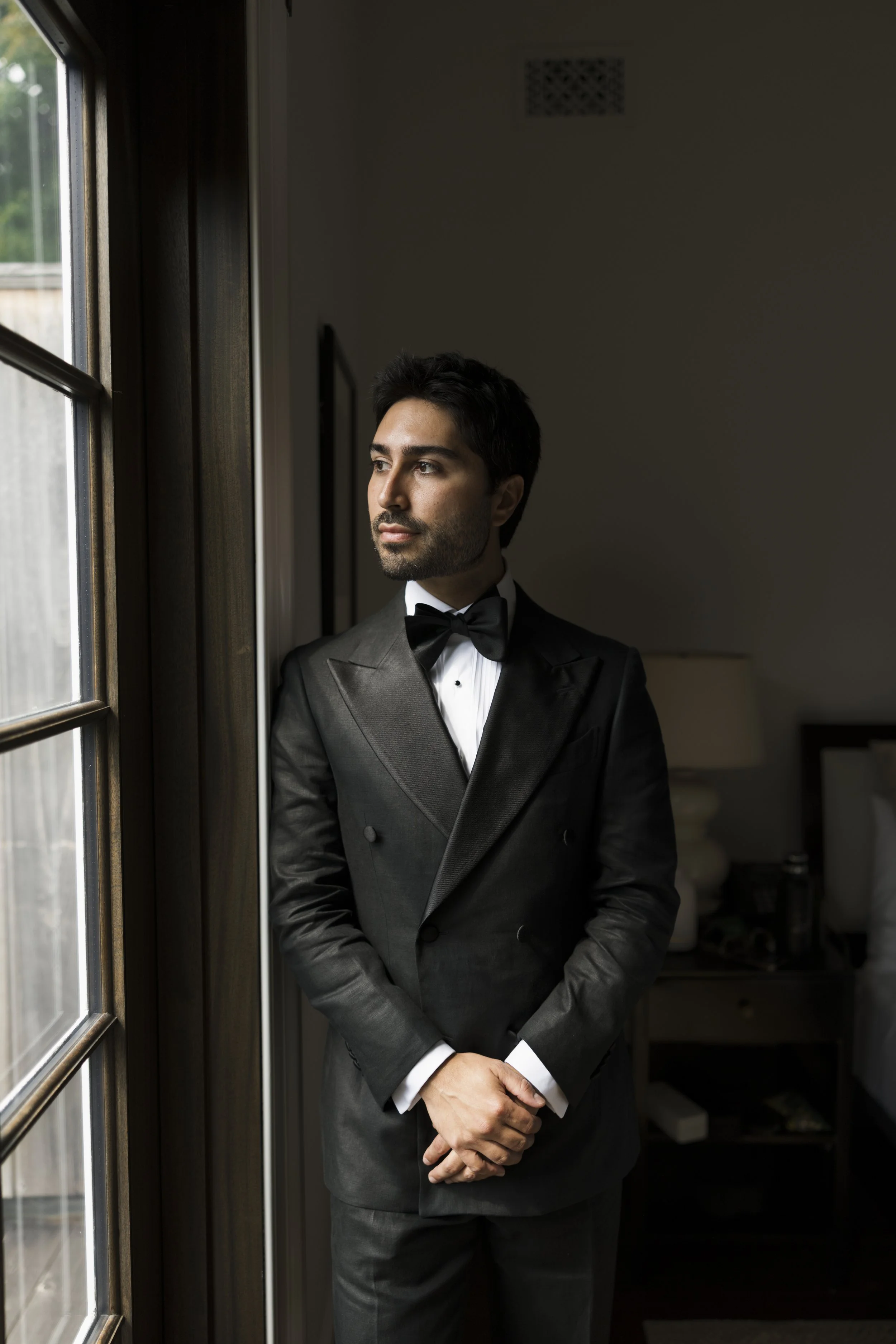 A man in a black tuxedo with a white shirt and black bow tie, standing indoors near a window, looking outside thoughtfully.