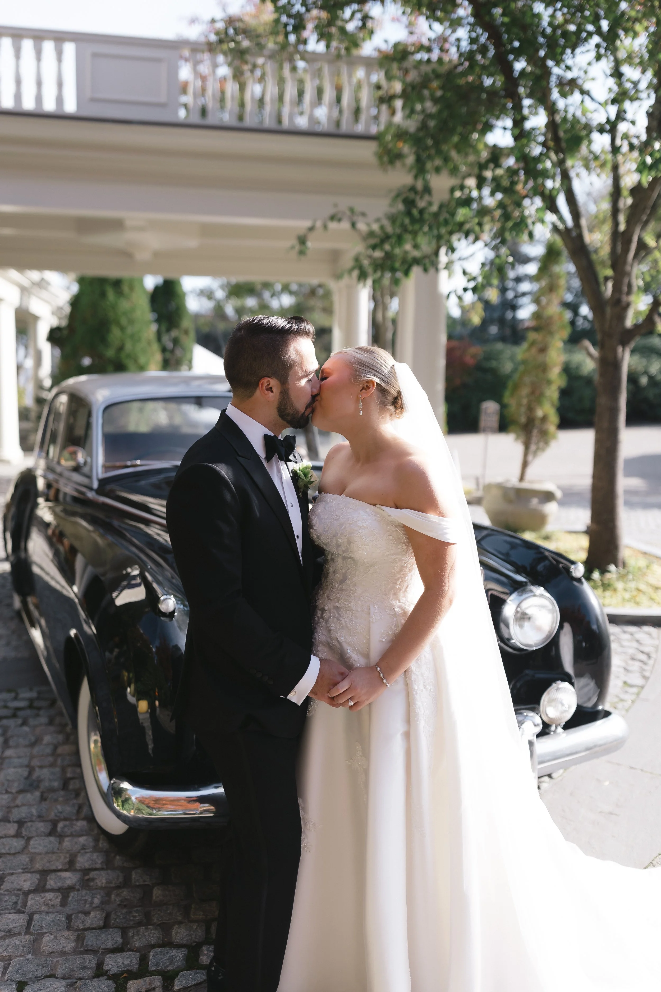 A bride and groom kiss in front of a vintage black car outdoors on their wedding day.