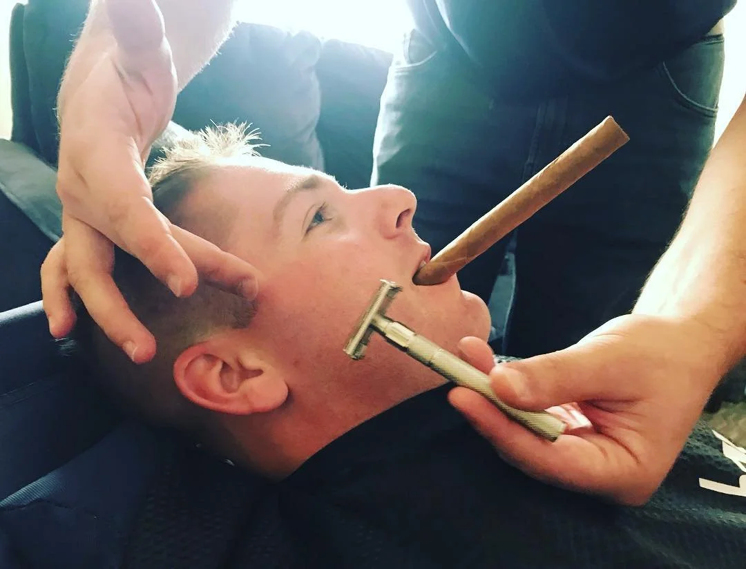 A man lying back with a cigar in his mouth being shaved with a straight razor as another person holds his face.