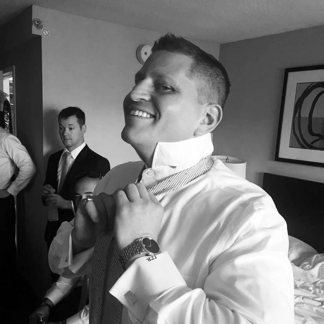 Man in a room adjusting tie, smiling, with other men in background.