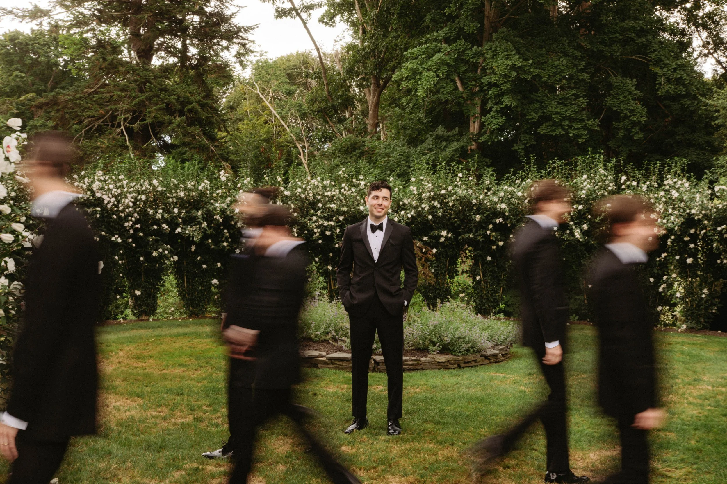 Man in a tuxedo standing in a garden with blurred people walking by