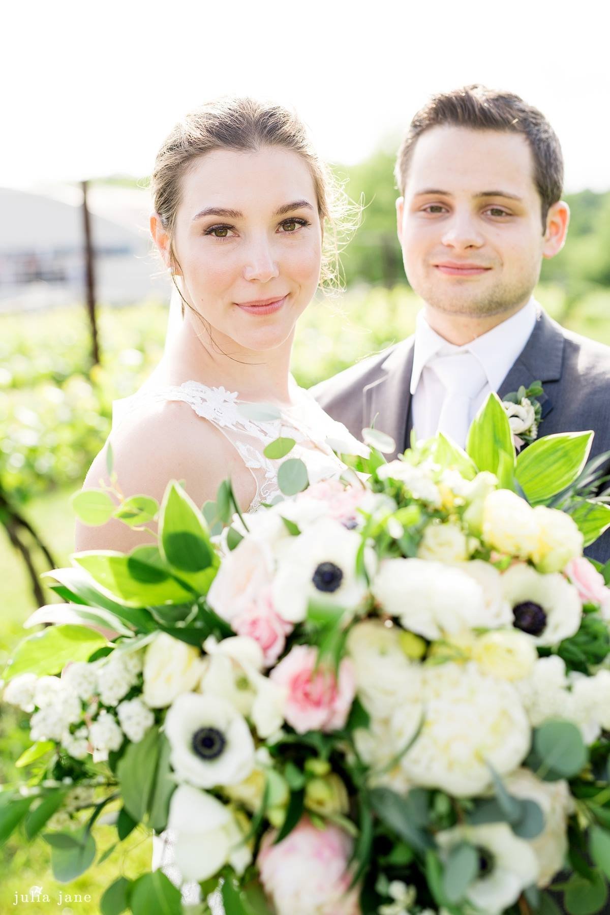 Bride and groom posing with a large bouquet of white and pink flowers outdoors.