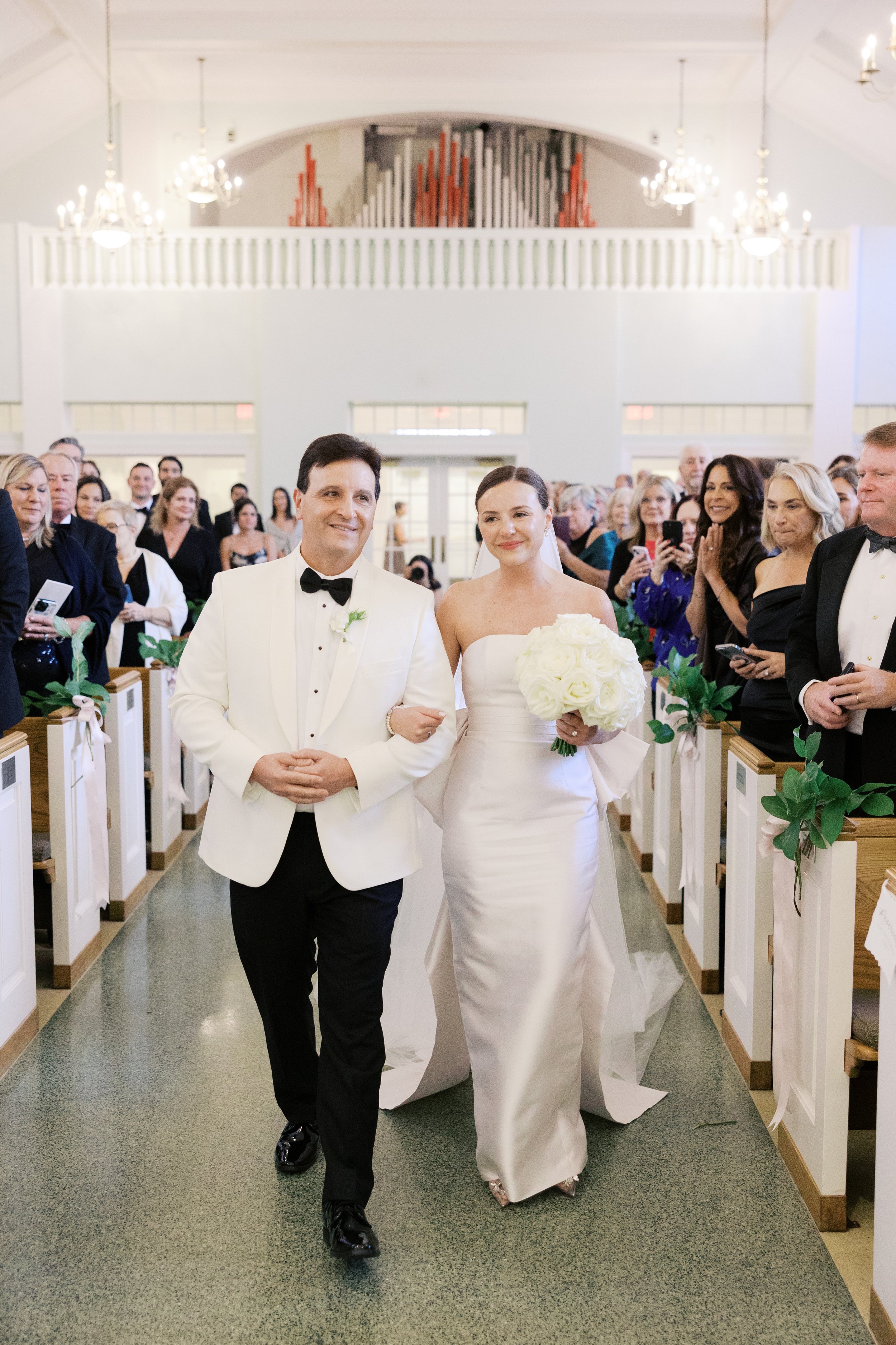 A bride holding a bouquet walks down an aisle in a church, accompanied by a man in a white tuxedo. The congregation is seated on both sides, observing the procession. The church is decorated with flowers and chandeliers.