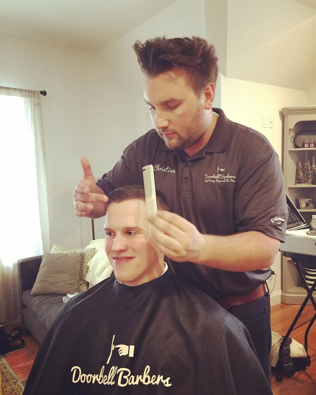 A barber giving a haircut to a smiling man in a cape with "Doorbell Barbers" printed on it, in a cozy room with furniture and soft lighting.