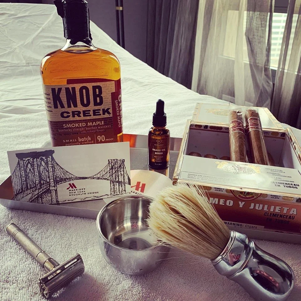 Collection of men's grooming and leisure items on a bed, including Knob Creek whiskey bottle, beard oil, shaving brush, metal shaving bowl, cigar box with cigars, safety razor, and hotel postcards.