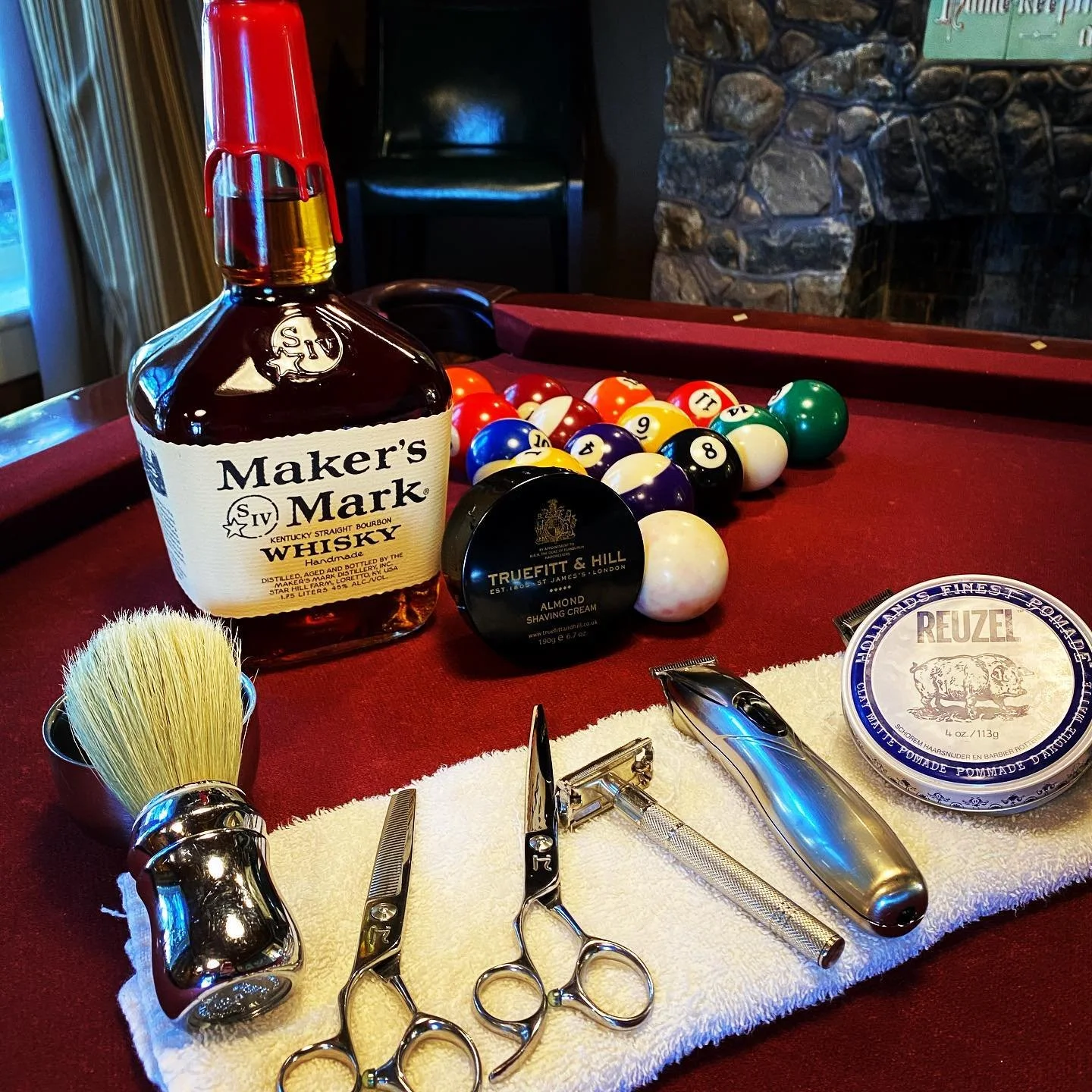 Barber tools and grooming products on a red pool table with a bottle of Maker's Mark whiskey and billiard balls in the background.
