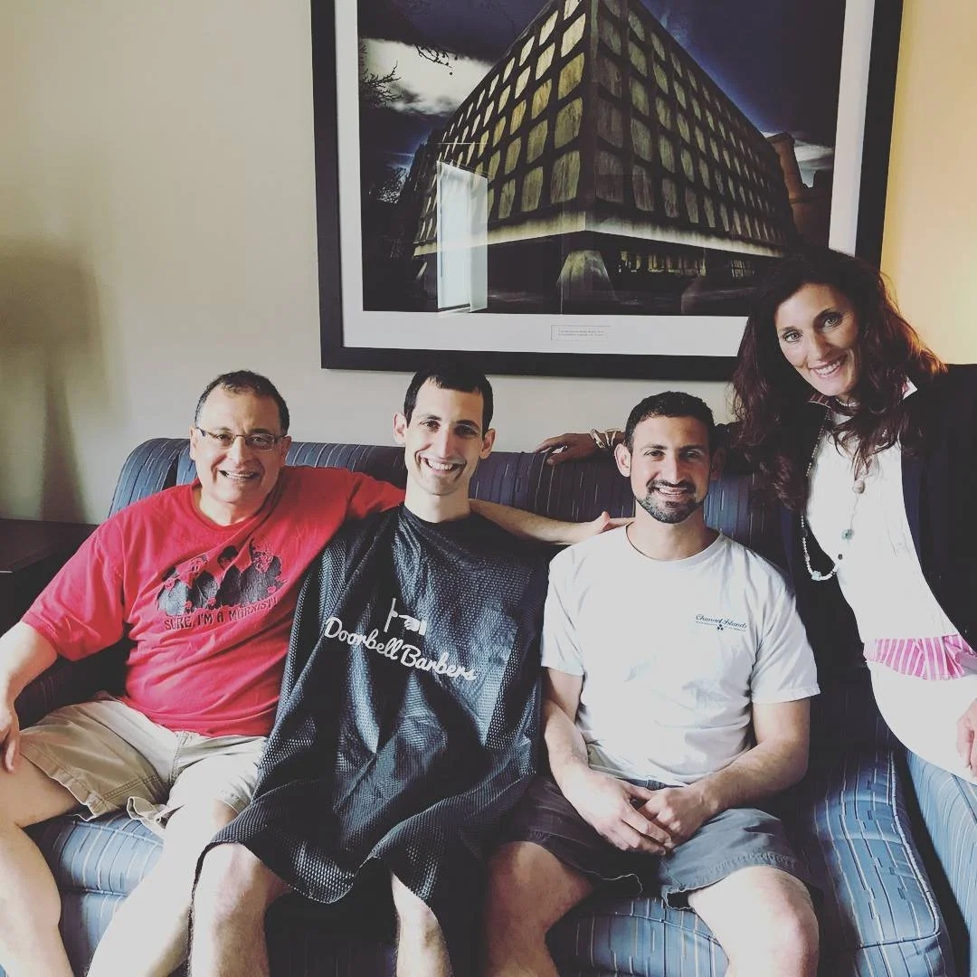 Four people sitting on a couch, three men and a woman, with a painting of a building on the wall behind them. One man is wearing a barber's cape.