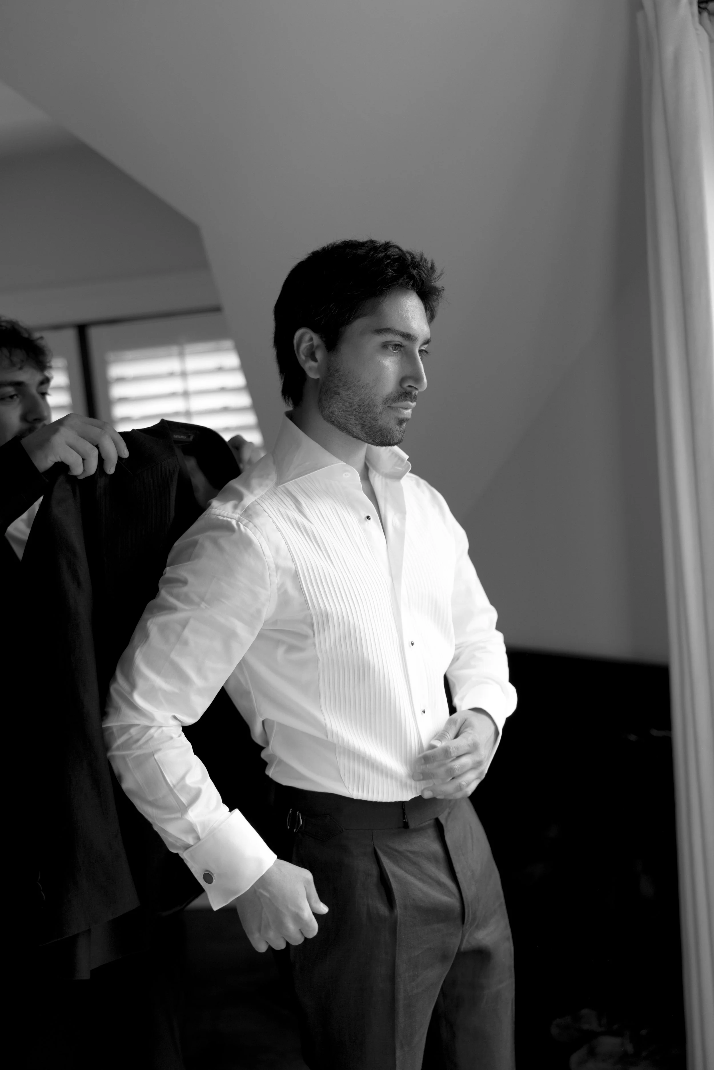 A man in a white dress shirt and dark trousers getting dressed, while a person behind him helps with a jacket, in a room with natural light.