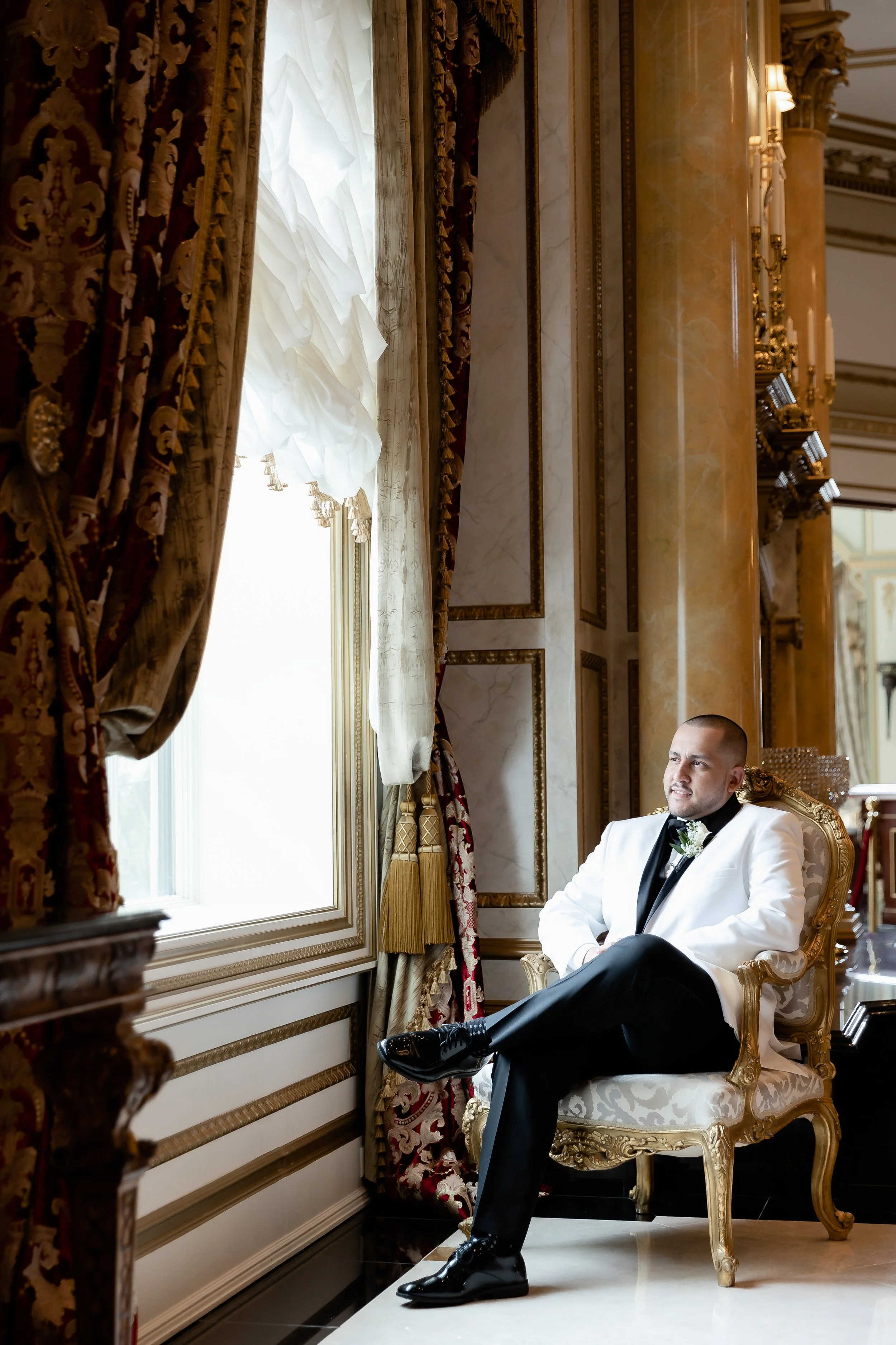 Man in white tuxedo sitting on ornate chair in elegant room with red and gold curtains, looking out a window.