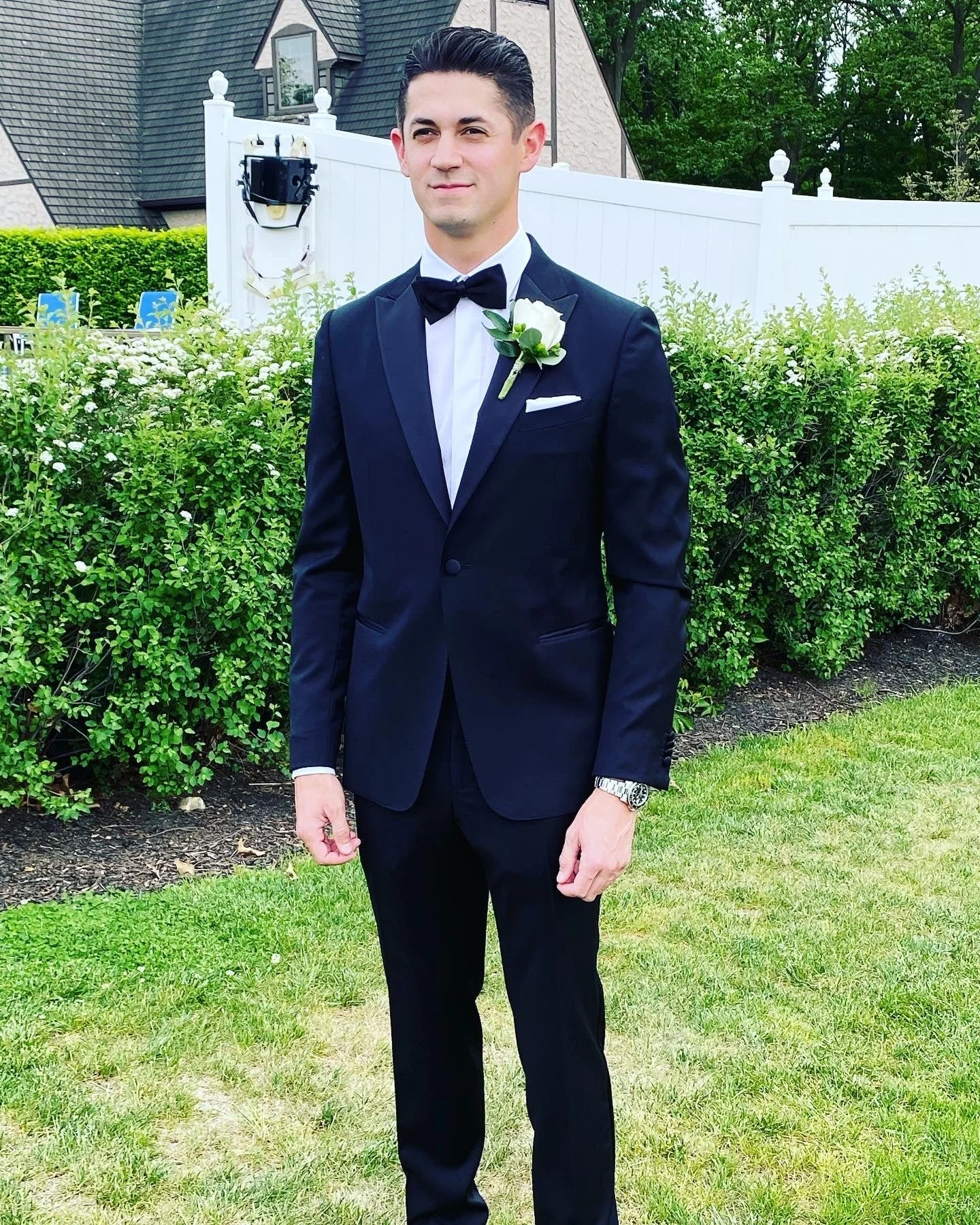 Man in a formal black tuxedo with a white shirt and bow tie, standing on grass with a white fence and shrubbery in the background.