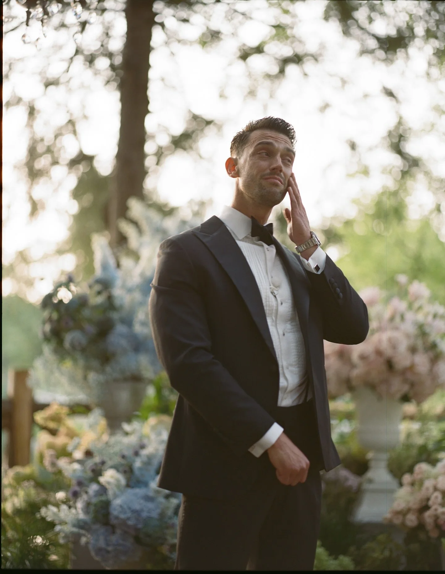 A man in a tuxedo standing outdoors at a wedding or formal event, holding his face with one hand, with flowers and trees in the background.