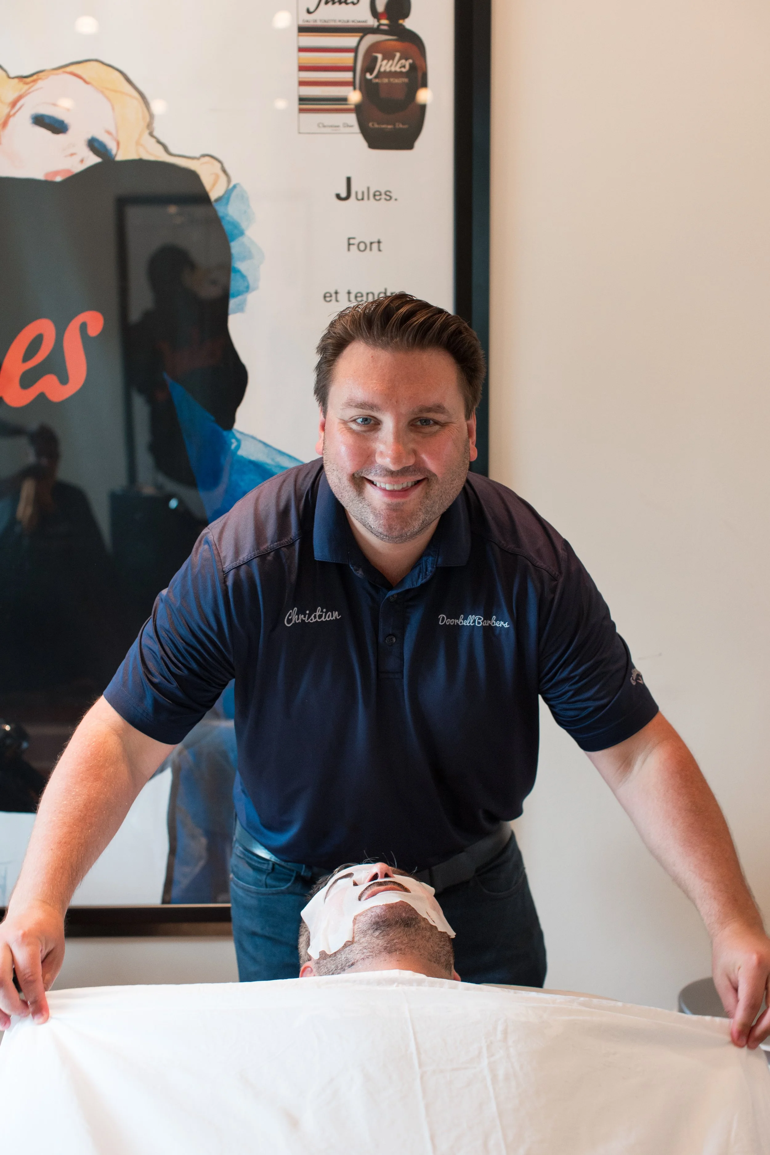 Barber standing over a client with a face mask in a barbershop, smiling, with a vintage poster in the background.