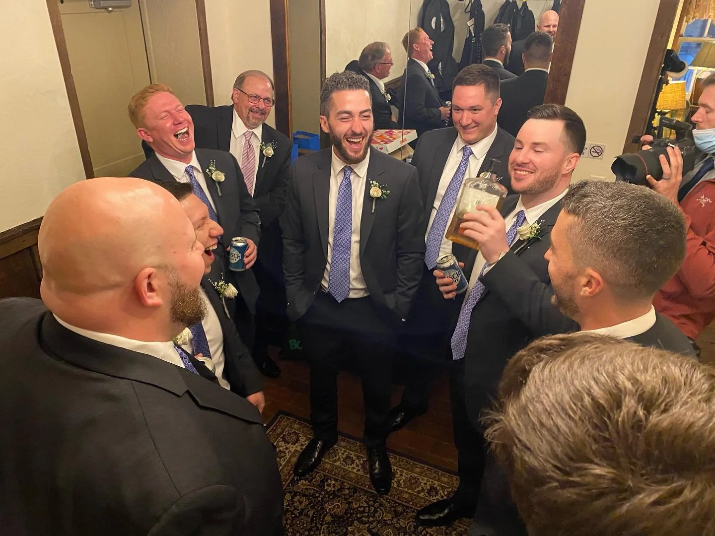 Group of men in suits, laughing and holding drinks, in a room with mirrors and wood paneling.