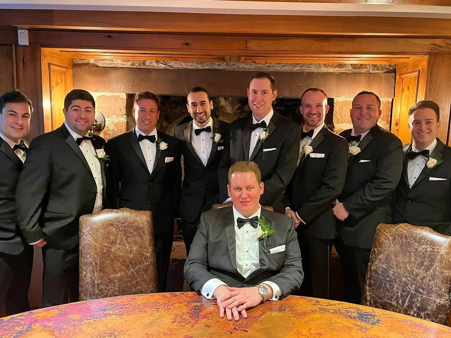 A group of men in tuxedos, one seated at a table, posing indoors. They are wearing black bow ties and white boutonnieres, suggesting a formal event like a wedding or celebration.