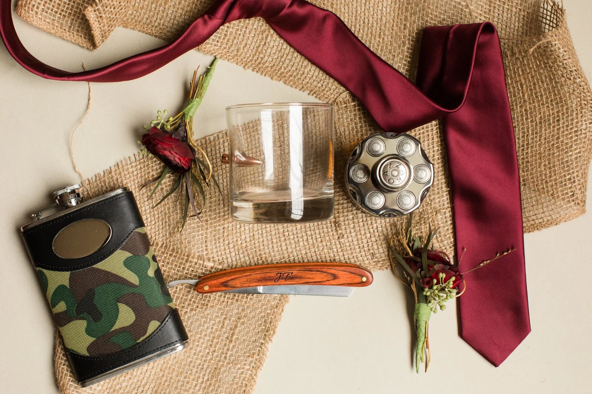 Men's gift set featuring a camouflage flask, a straight razor, a bullet whiskey glass, burgundy tie, boutonnieres, and a round hip flask, arranged on burlap fabric.