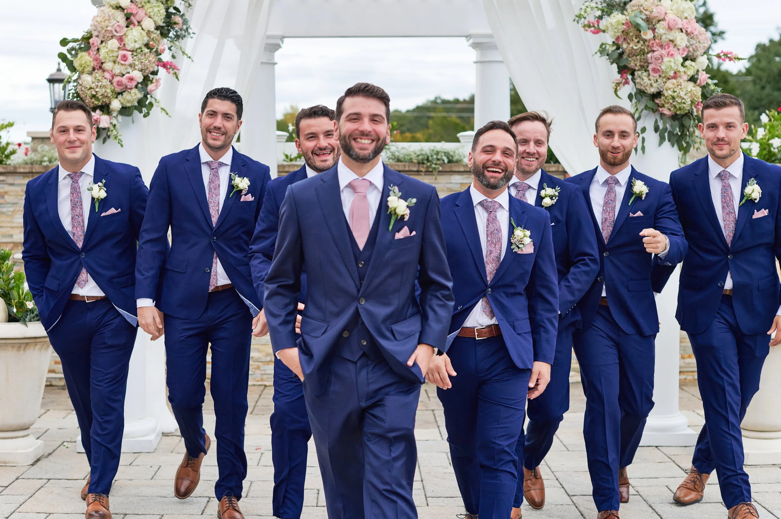 Groom and groomsmen in blue suits with floral ties and boutonnieres, walking under an arch with floral decorations.