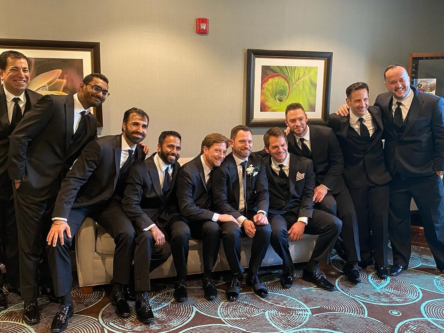 Group of men in formal attire posing indoors, some sitting on a couch, others standing behind, with framed pictures on the wall.