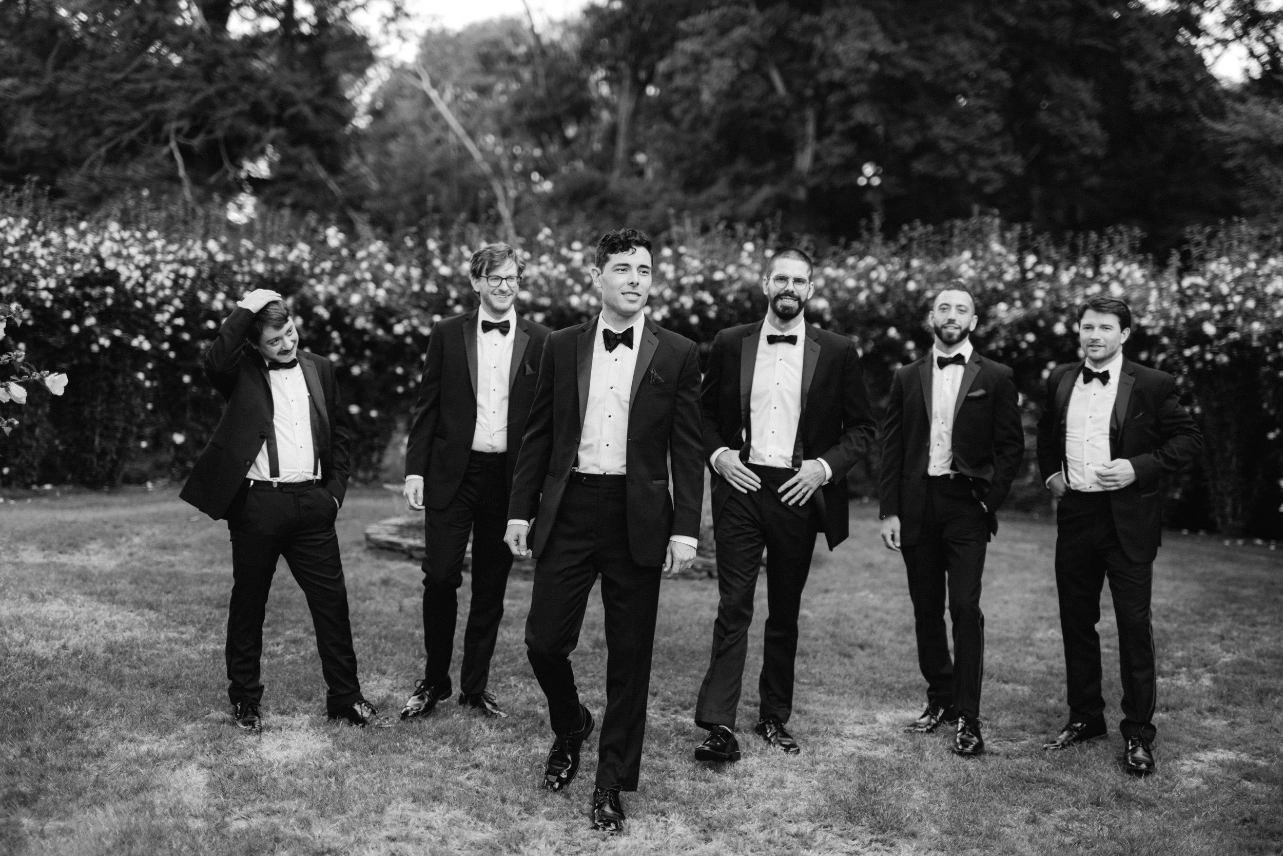 Black and white photo of six men in tuxedos standing outdoors in a garden setting with trees and flowers in the background.
