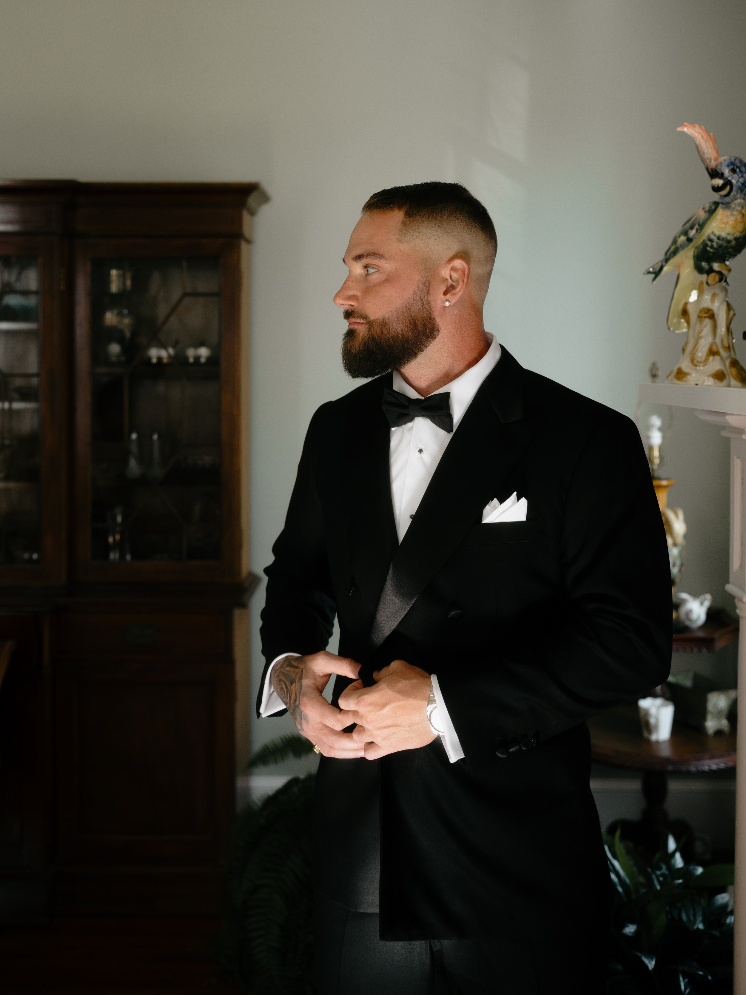 A man dressed in a black tuxedo with a bow tie, standing indoors with a serious expression, adjusting his jacket.