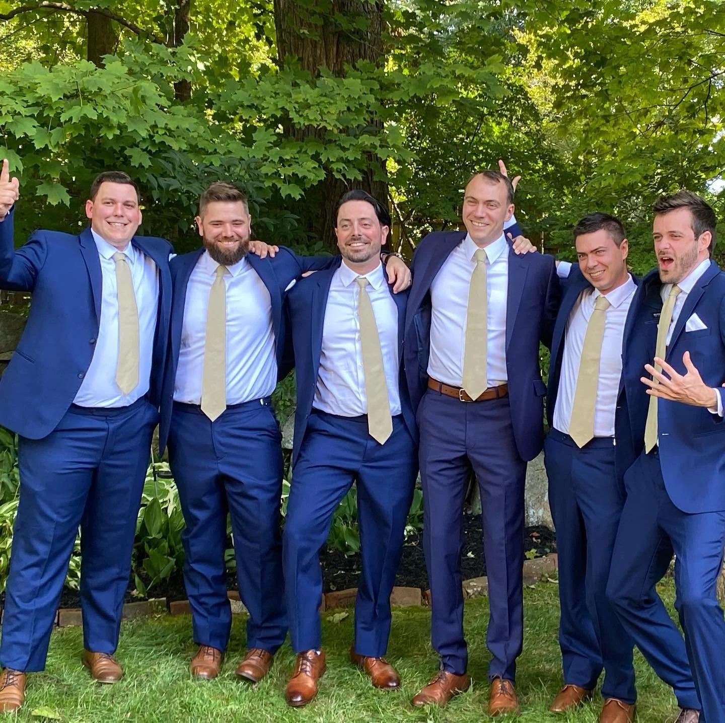 Group of six men wearing matching blue suits and beige ties, standing together and smiling in an outdoor garden setting with greenery in the background.