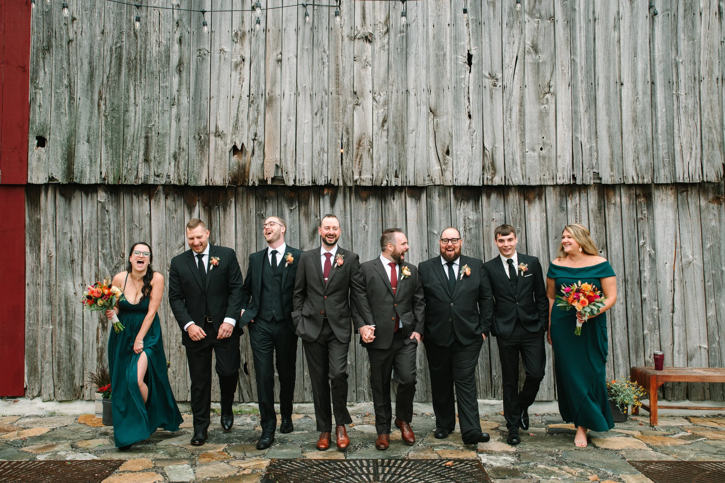Group of wedding party members in formal attire walking and laughing in front of a rustic wooden barn.
