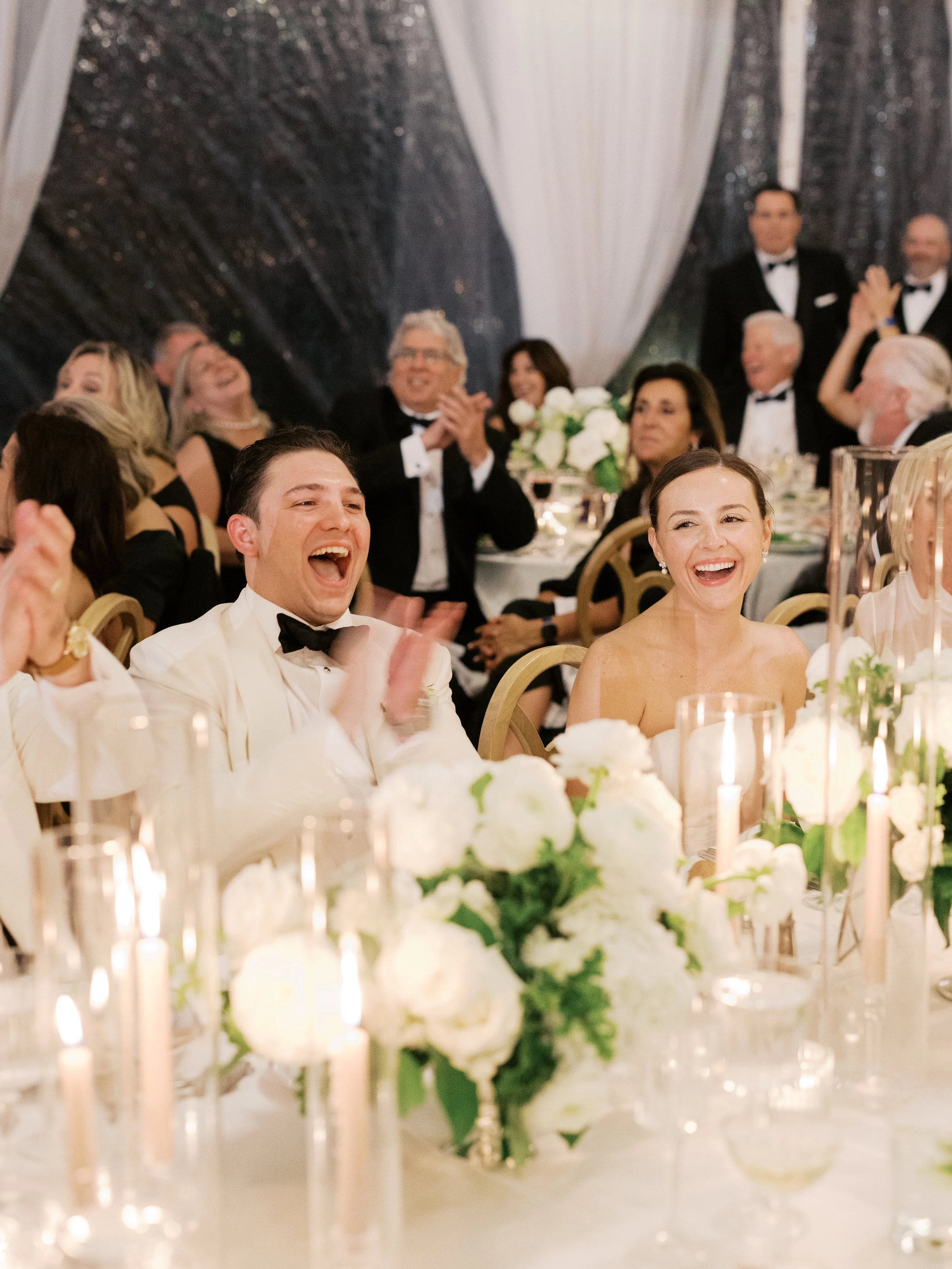 A couple seated at a wedding reception table, smiling and laughing, with floral arrangements and candles in the foreground. Guests in formal attire are visible in the background, clapping and enjoying the event.
