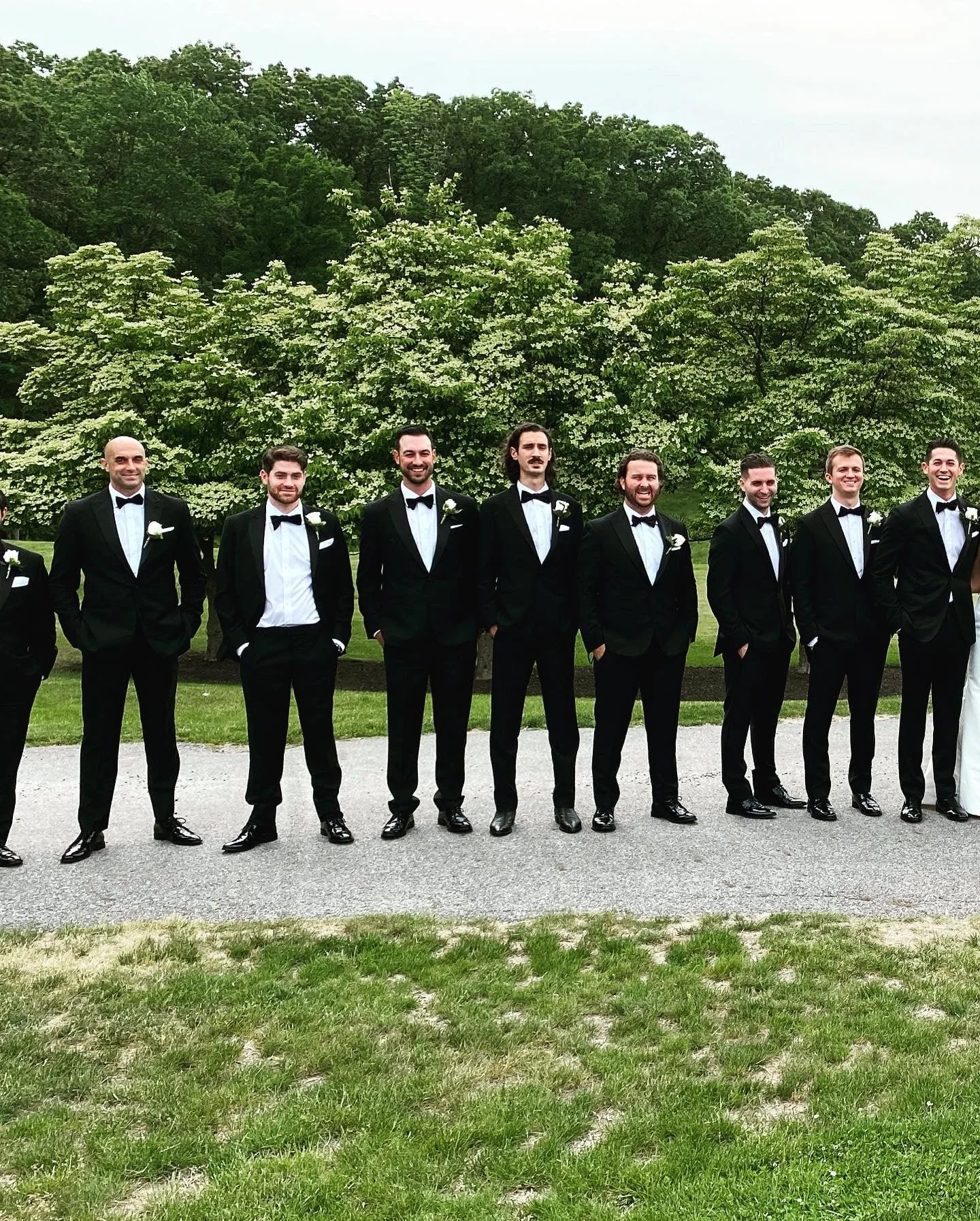 Group of men in tuxedos posing outdoors with greenery and trees in the background.