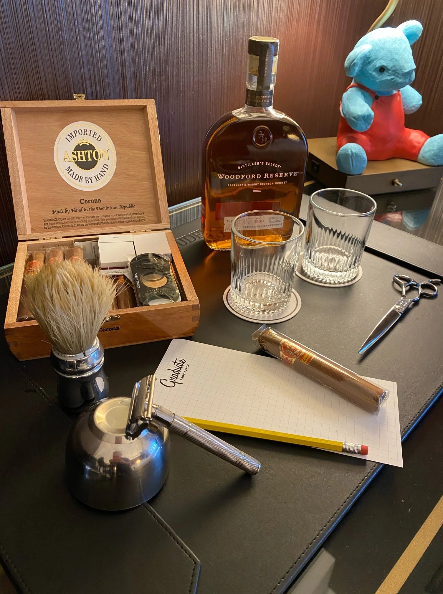 Assorted items on a desk, including a bottle of Woodford Reserve bourbon, two glasses, a wooden cigar box labeled 'Ashton,' shaving brush and razor, scissors, a notepad labeled 'Gravati,''' a pencil, a single cigar, and a small blue teddy bear wearin